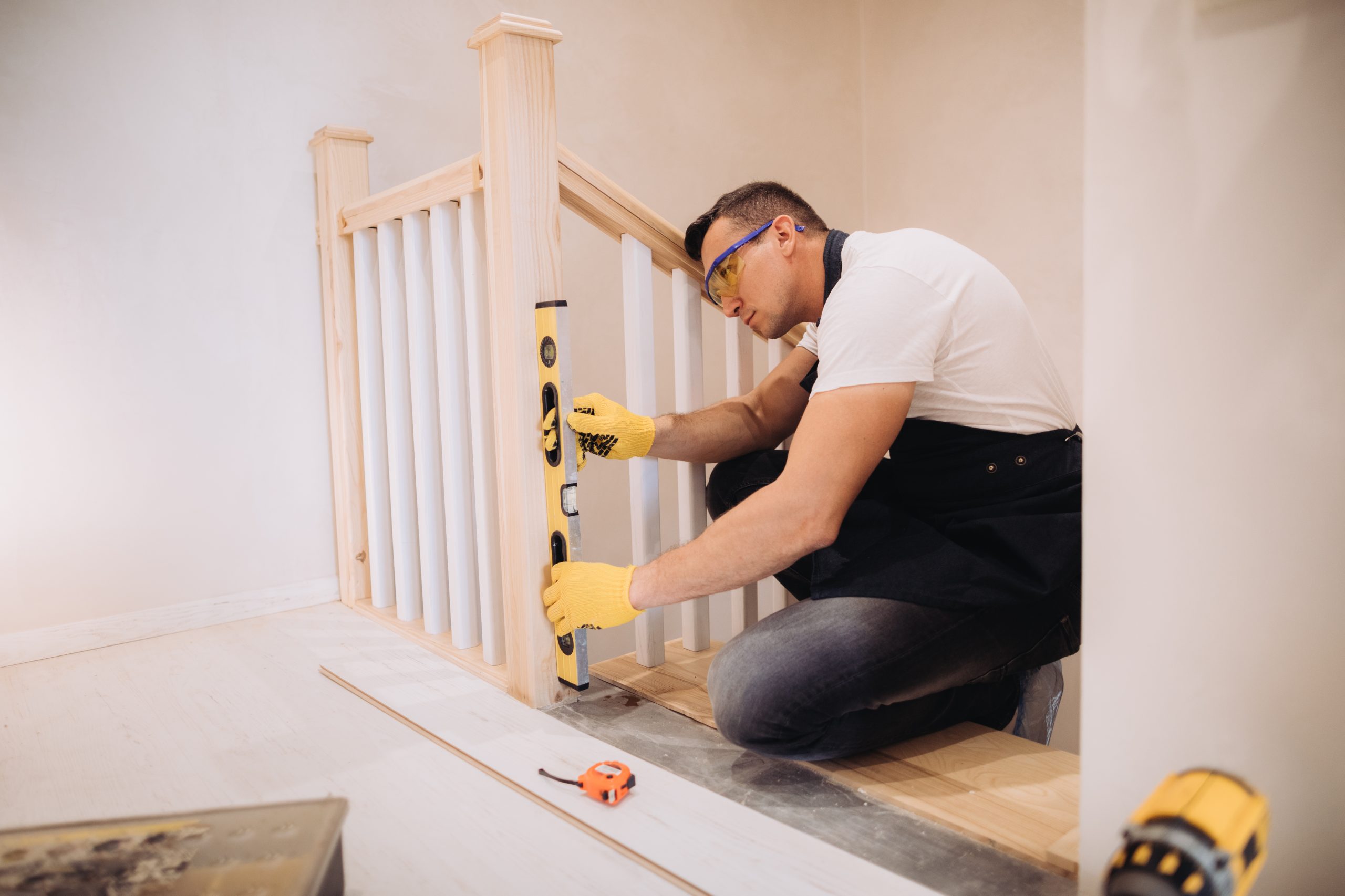 Maintenance man is working with a level measurement tool near the staircase. He is wearing glasses, yellow gloves and a black apron